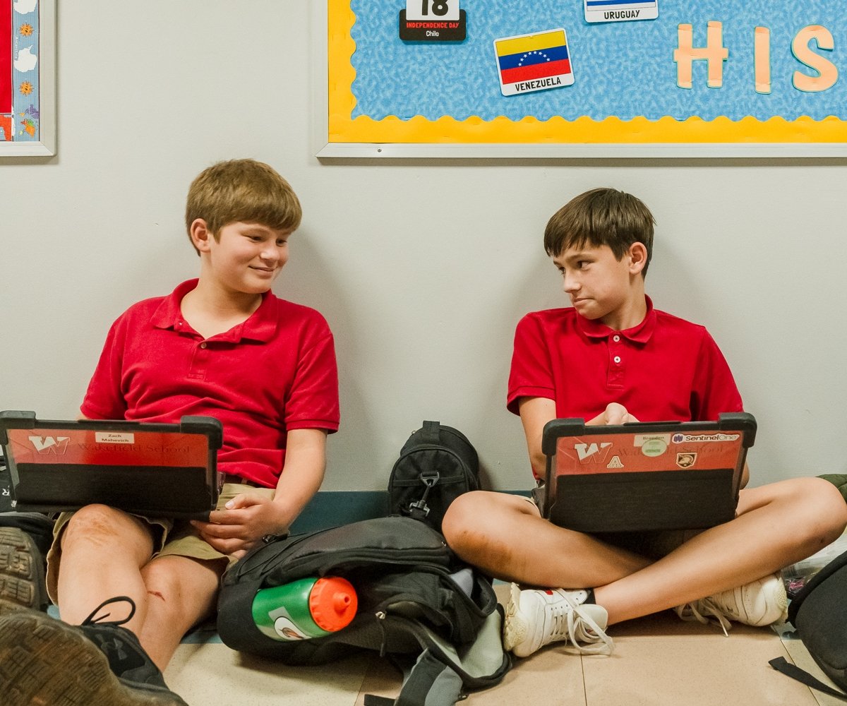 two middle school students sitting in the hallway working on laptops